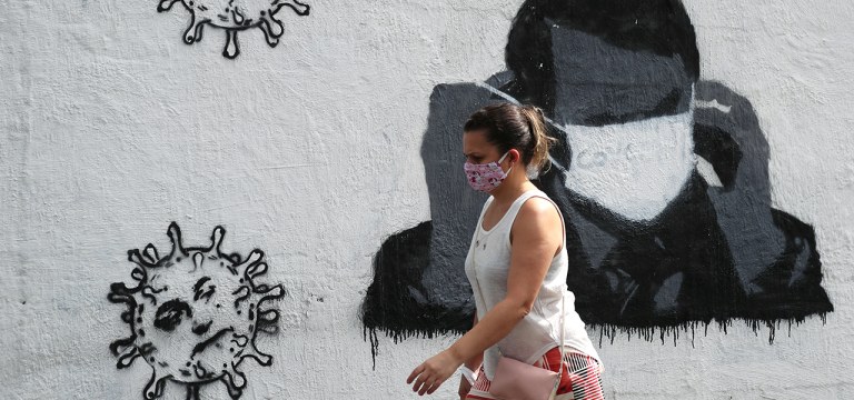 Mulher passa por grafite do presidente Jair Bolsonaro ajustando sua máscara protetora e em forma de vírus, em meio ao surto de Covid-19 no Rio de Janeiro.